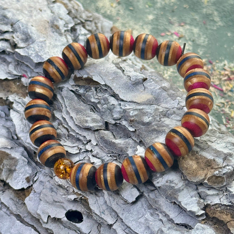 Bracelet made from recycled skateboard wood with pink, blue and black colour tones, round hand-shaped beads on elastic cord and one natural amber bead, shown on textured bark surface.