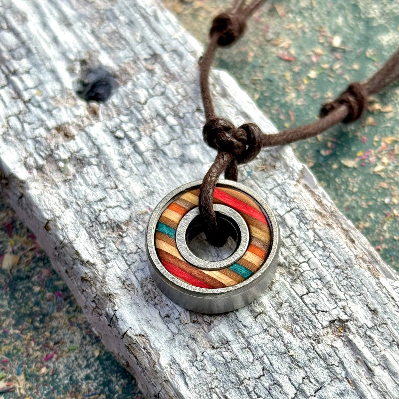 A small round ball-bearing pendant with red, blue and orange skateboard wood colour tones, shown on a brown adjustable cord on a textured wooden surface.