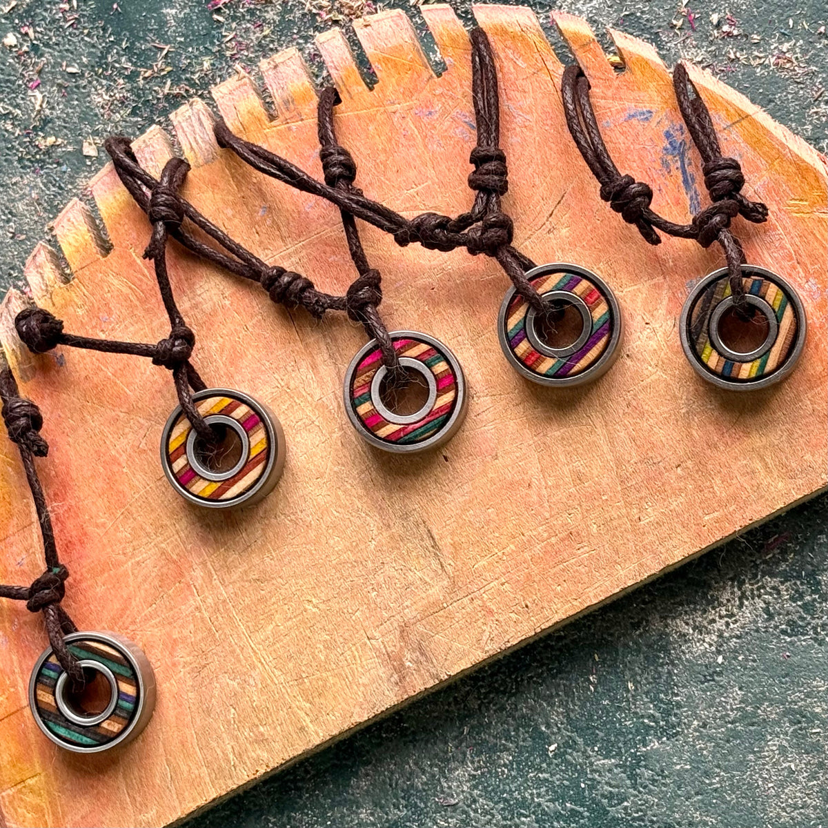 Five recycled skateboard pendant necklaces on brown cords, each with a steel skateboard bearing set into layered maple, laid on a cut skateboard tail.