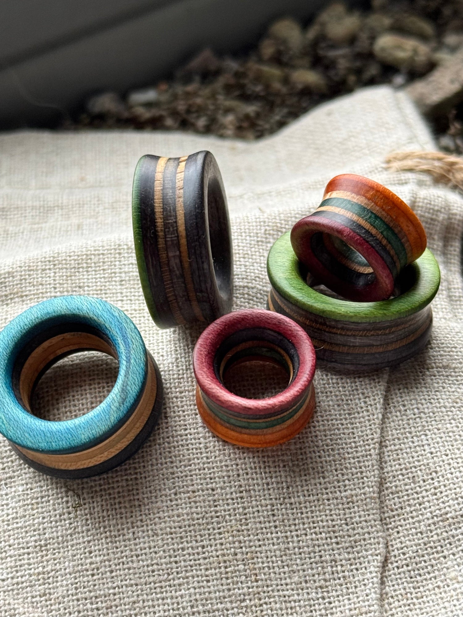 A colourful stack of wooden ear tunnels made from recycled skateboards, featuring layered tones of teal, red, green, black, and natural wood grain, displayed on a neutral fabric background.