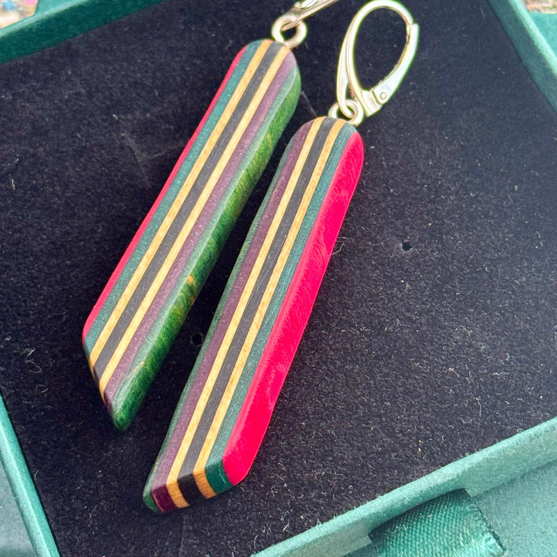 Woman wearing long pink earrings made from recycled skateboard wood, standing outside on a sunny day