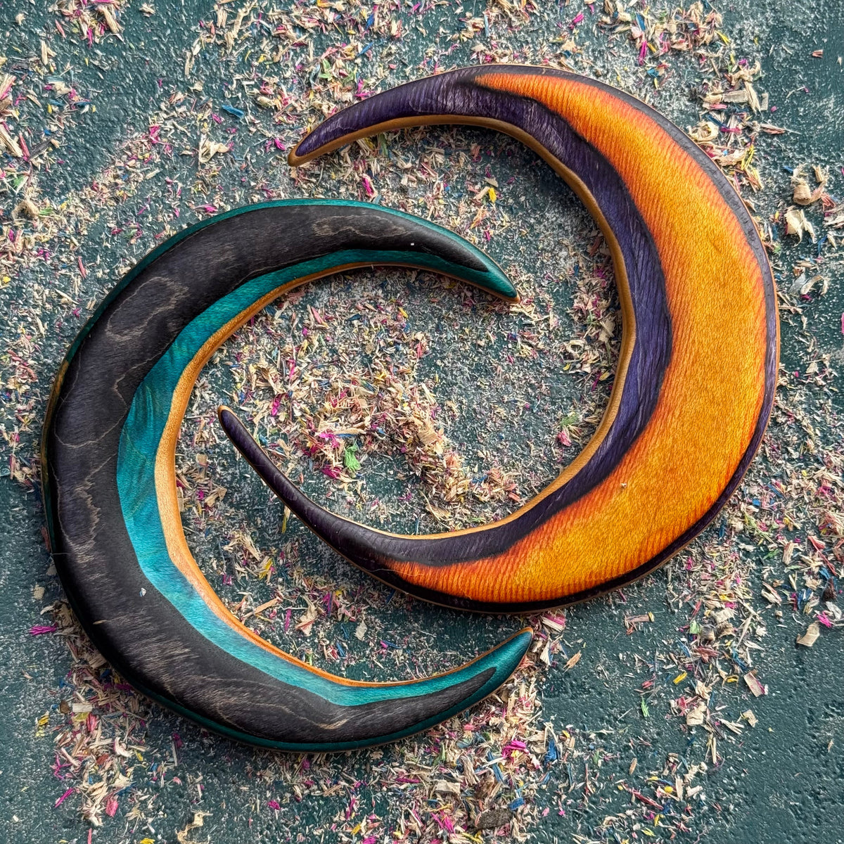 Two crescent-shaped hair forks made from recycled skateboard wood, one with teal and black layers and the other with orange and purple grain, resting on a green surface covered in colourful wood shavings.