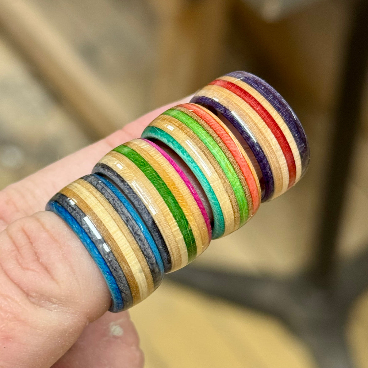 Four recycled skateboard wood rings worn stacked on one finger, showing blue, grey, green, pink, teal, orange, red and purple maple layers.