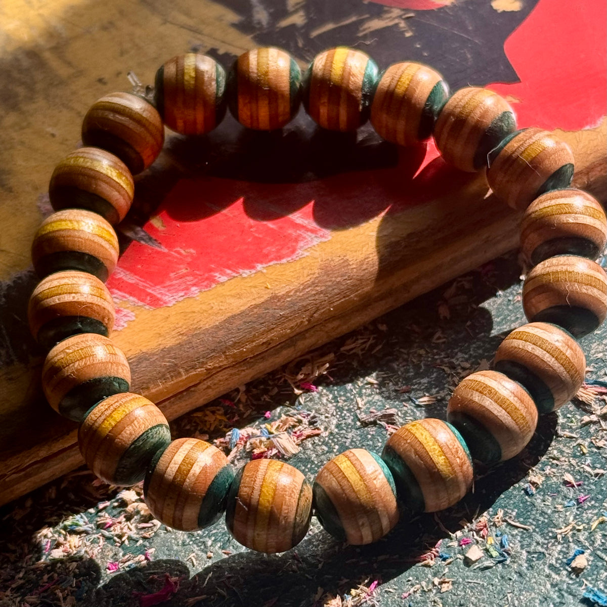 Light teal and yellow striped skateboard maple beads, bracelet resting across a red skateboard deck in sunlight.