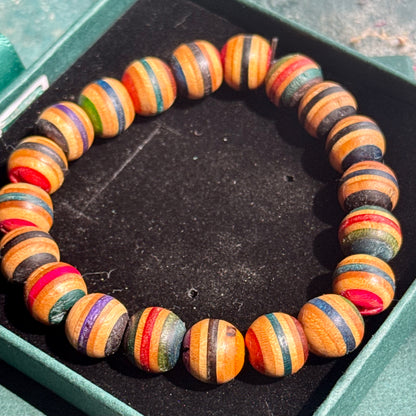 Assorted Colour Skateboard Bracelet featuring round wooden beads with multicolored stripes, displayed in a green box on a black fabric background.