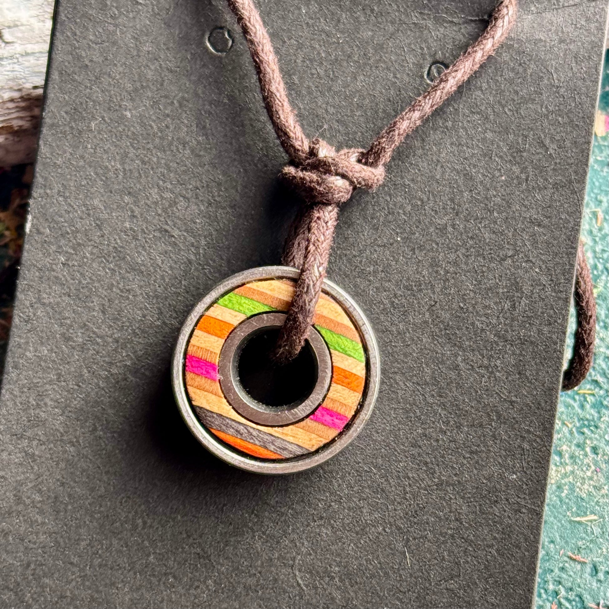 Skateboard bearing pendant with orange, pink and green recycled maple layers on a brown waxed cord.