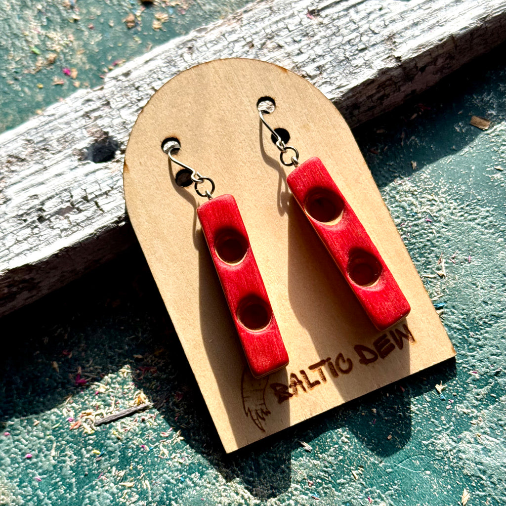 Red recycled skateboard wood bar earrings with carved round hollows, resting on weathered wood
