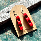 Red recycled skateboard wood bar earrings with carved round hollows, resting on weathered wood