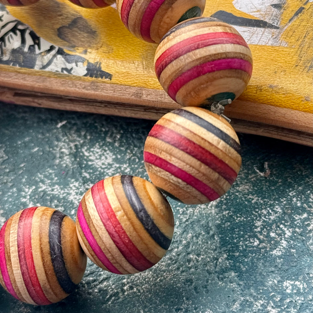 Chunky wooden beads with red, pink, black and teal layers from a recycled skateboard deck, on a yellow deck edge.