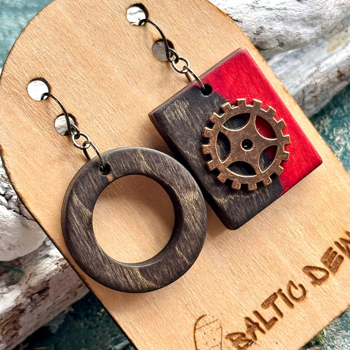 Close view of a charcoal wooden ring and red square earring with a decorative brass cog