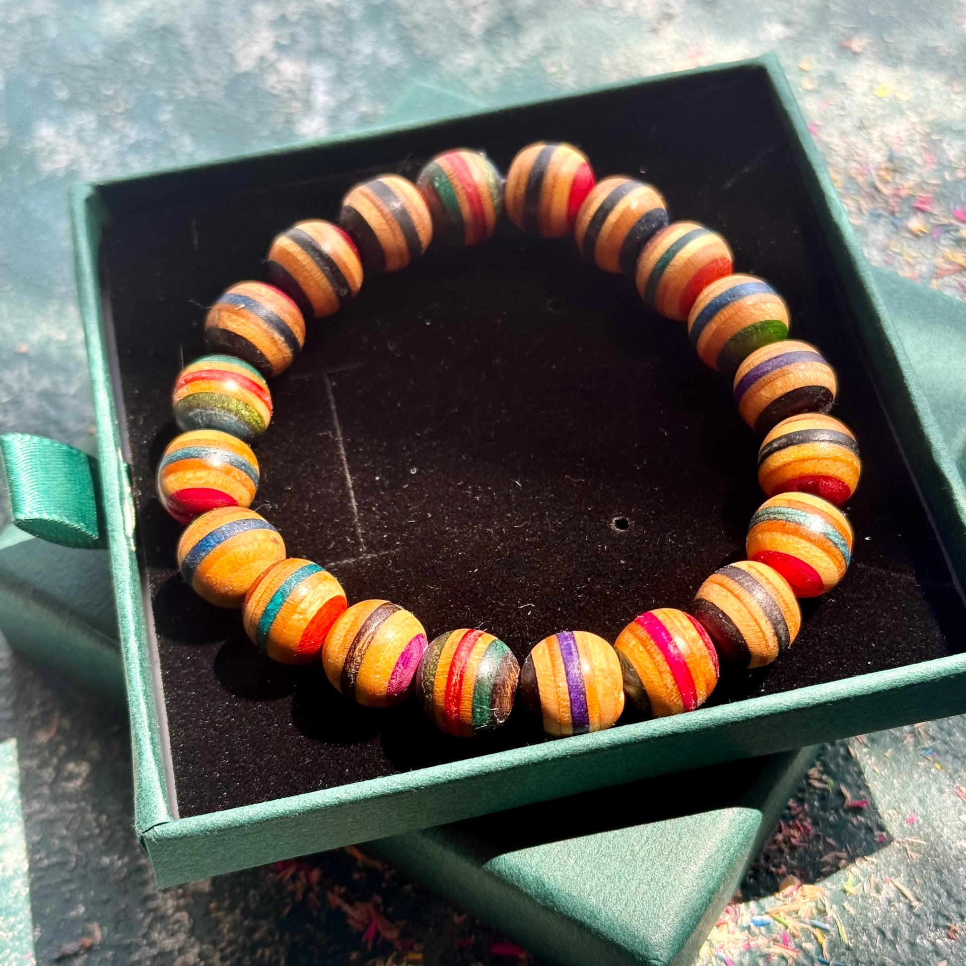 Assorted Colour Skateboard Bracelet with round, striped wooden beads in multiple vibrant colors displayed in a green box on a textured surface.