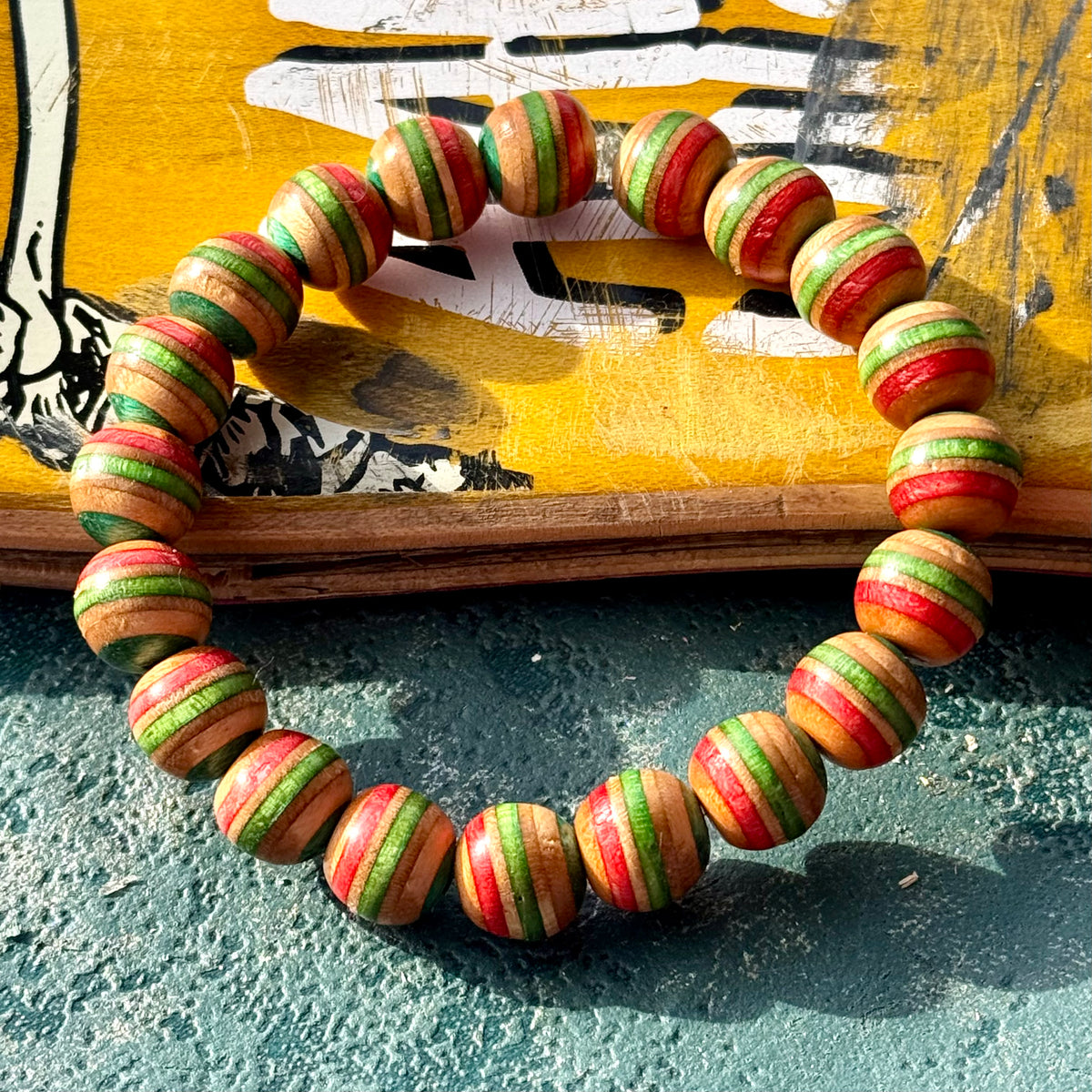 Red, green and teal wood bead bracelet from recycled skateboard maple in a full round on a yellow deck.
