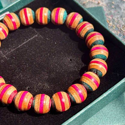 Close-up of a Pink and Teal Skateboard Bracelet made of round beads with alternating pink and teal stripes on a wooden texture, displayed in a green box.