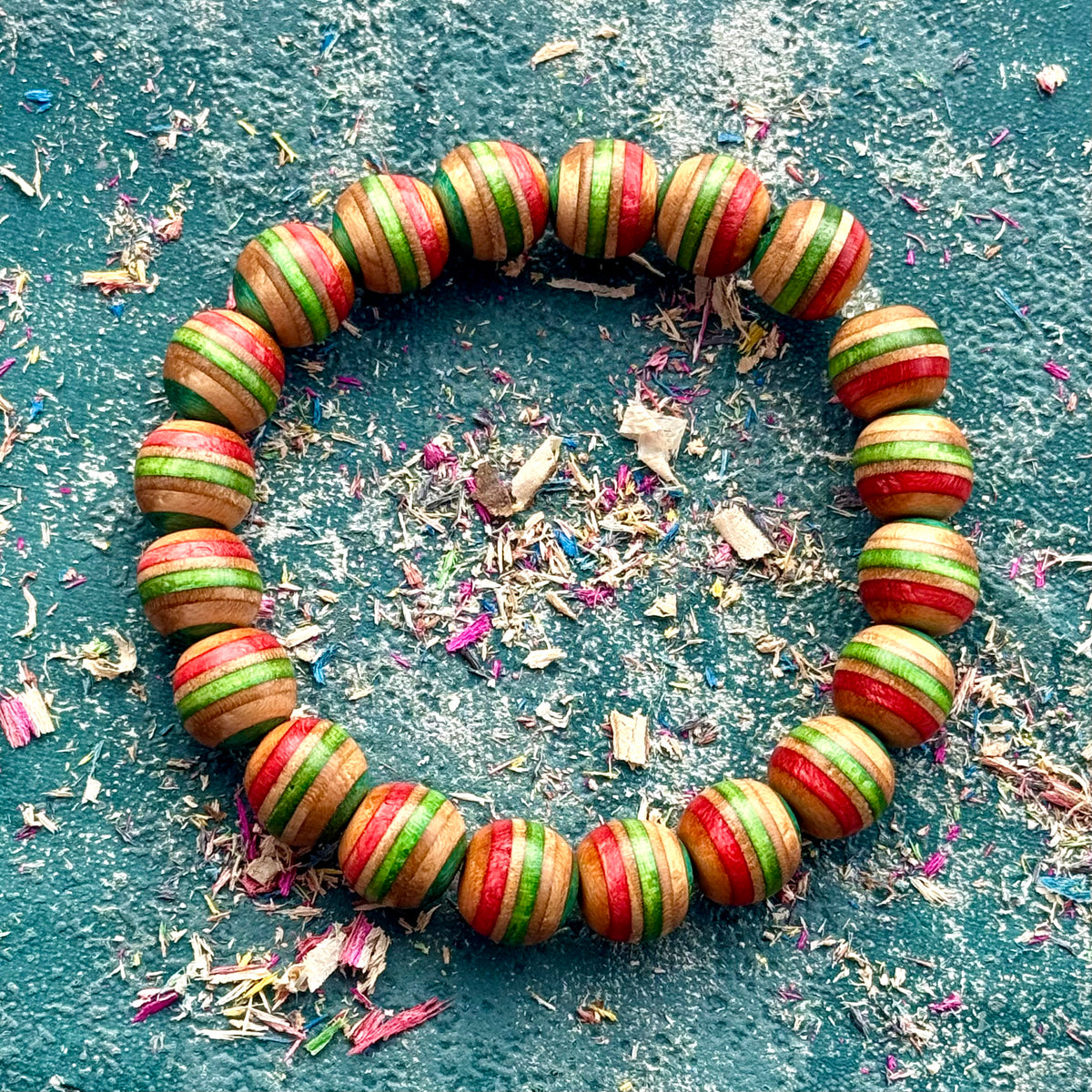 Red, green and teal wooden bead bracelet seen from above among wood shavings on a dark surface.