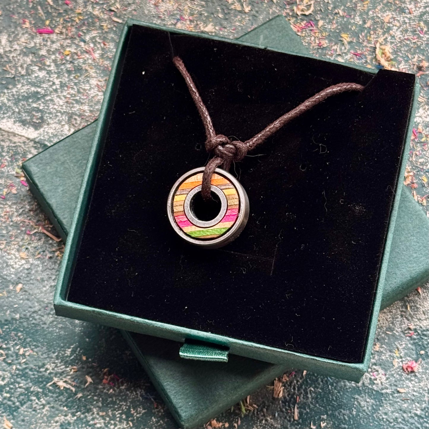 Skateboard bearing pendant necklace displayed in an open green jewellery box, showing pink, yellow and green maple layers.
