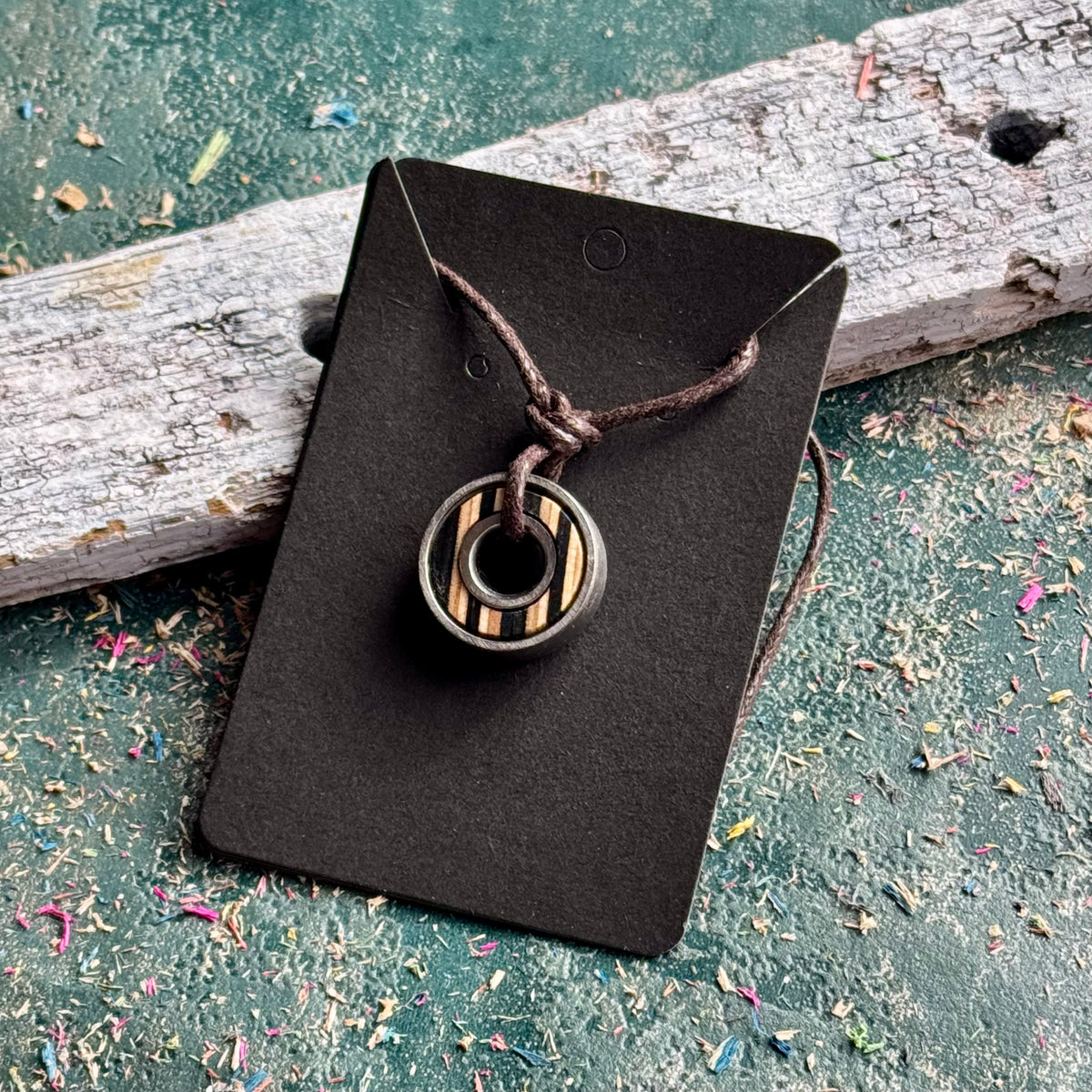 Skateboard bearing pendant necklace with black and natural maple stripes, shown on a Baltic Dew card.