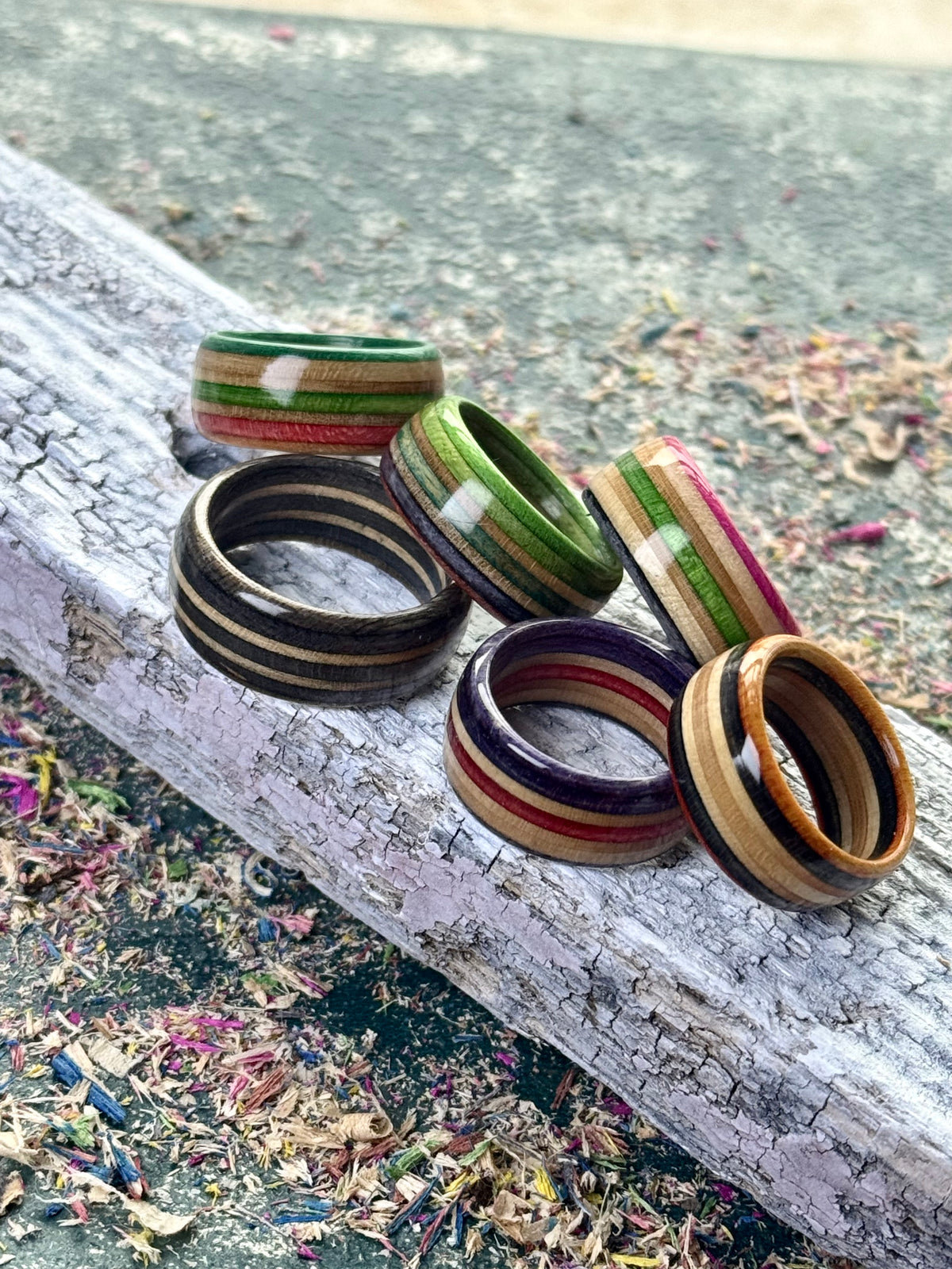 Five recycled skateboard wood rings resting on weathered timber, with green, pink, purple and red layers showing.