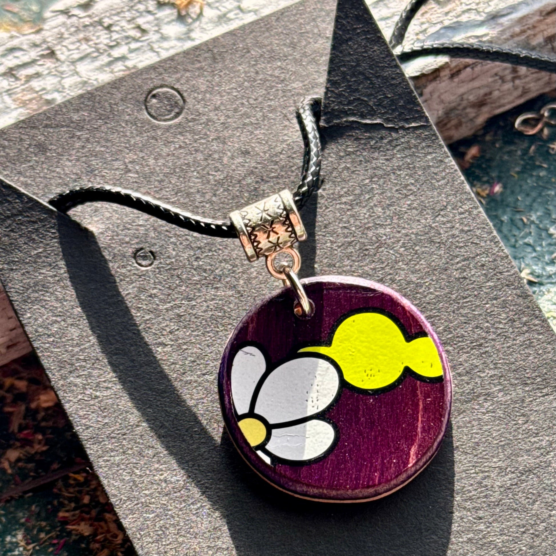 Round purple wooden pendant showing the skateboard deck's white flower graphic on a black waxed cord.