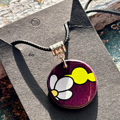 Round purple wooden pendant showing the skateboard deck's white flower graphic on a black waxed cord.
