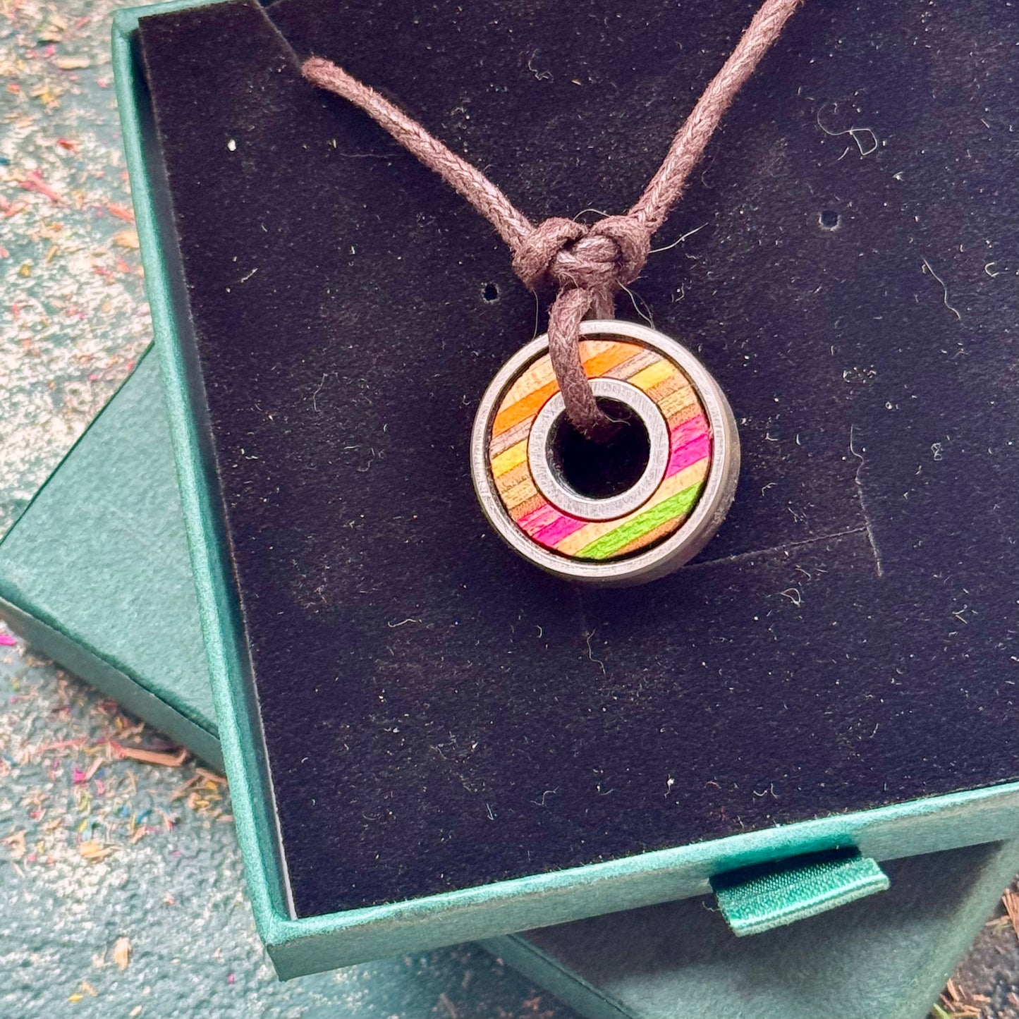 Skateboard bearing pendant necklace with pink, yellow and natural maple layers displayed on a green jewellery box.
