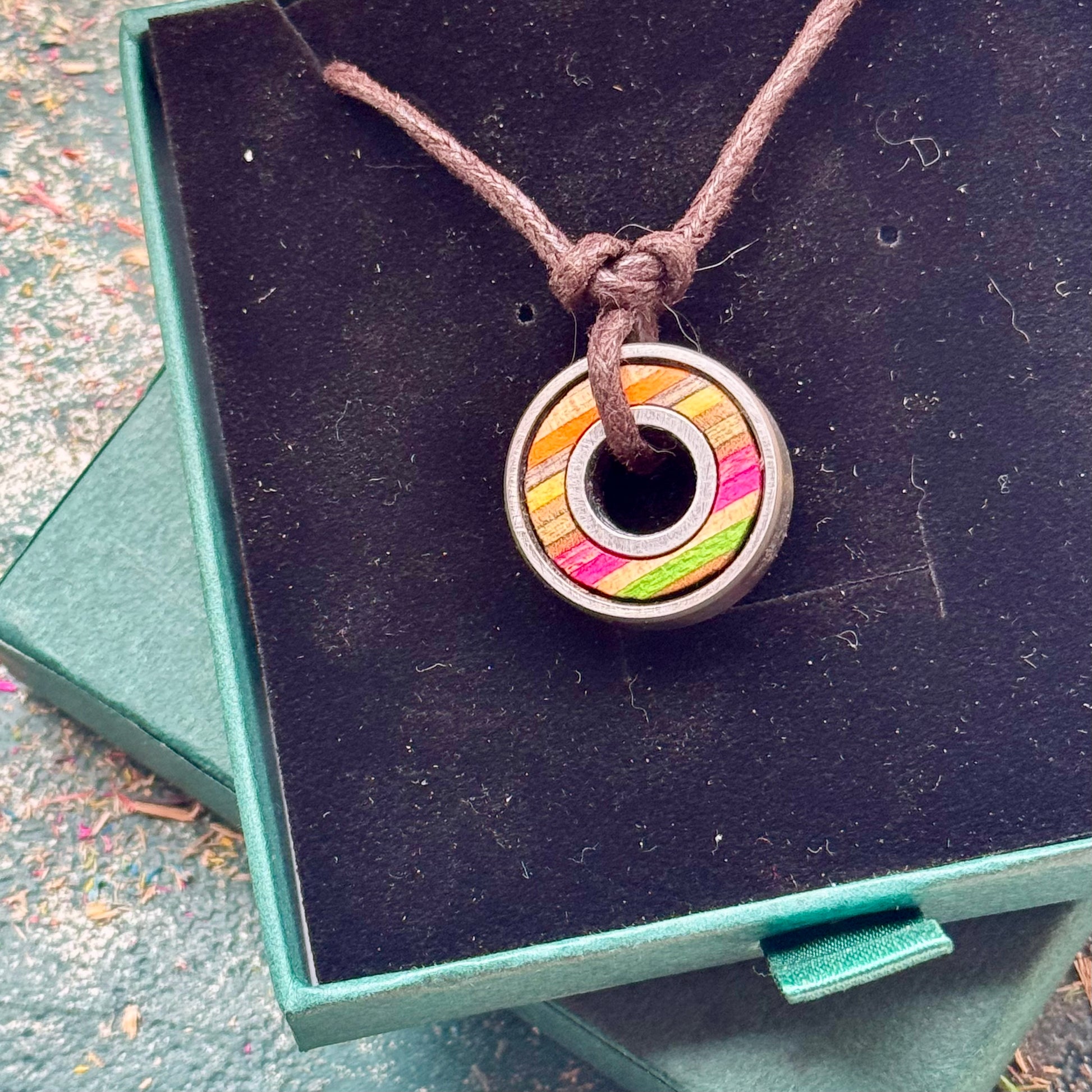 Skateboard bearing pendant necklace with pink, yellow and natural maple layers displayed on a green jewellery box.