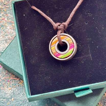 Skateboard bearing pendant necklace with pink, yellow and natural maple layers displayed on a green jewellery box.