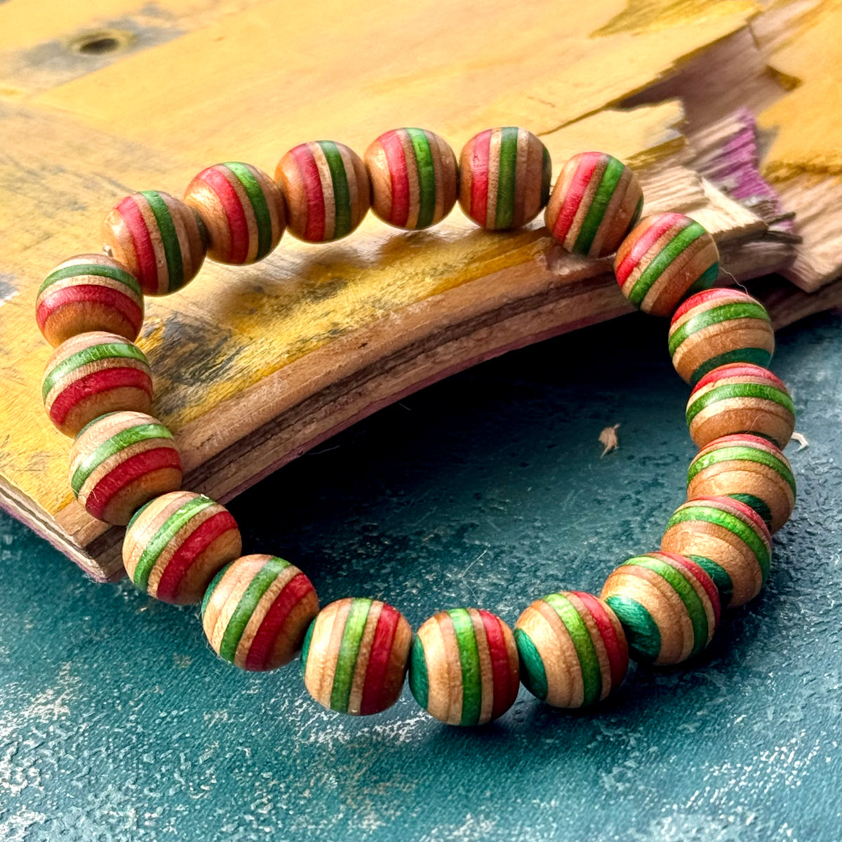 Red, green and teal skateboard wood bead bracelet laid on broken deck layers, handmade by Baltic Dew.