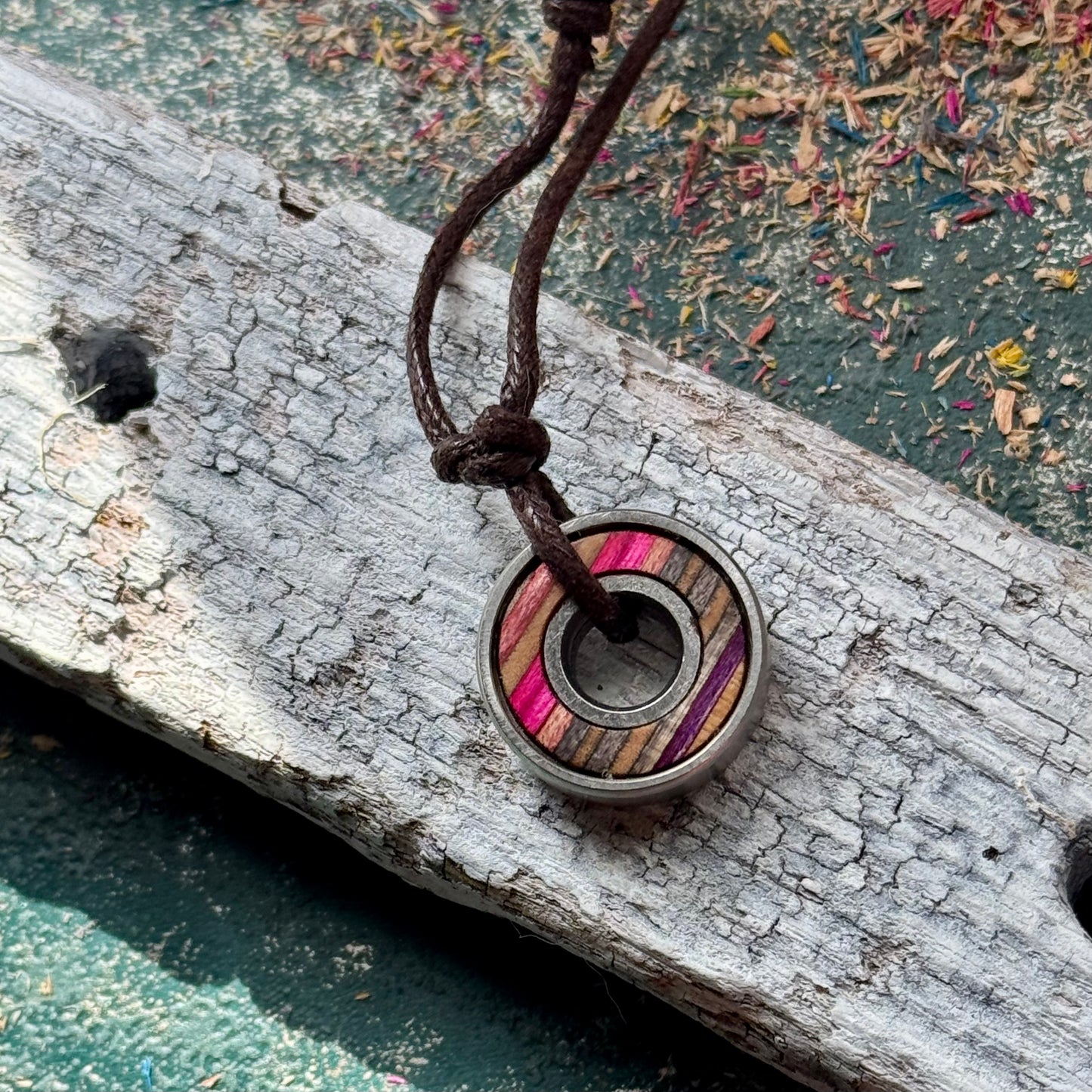 Skateboard bearing pendant necklace with pink and grey maple layers resting on weathered wood.