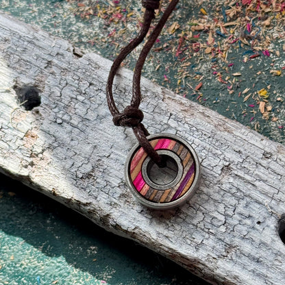 Skateboard bearing pendant necklace with pink and grey maple layers resting on weathered wood.