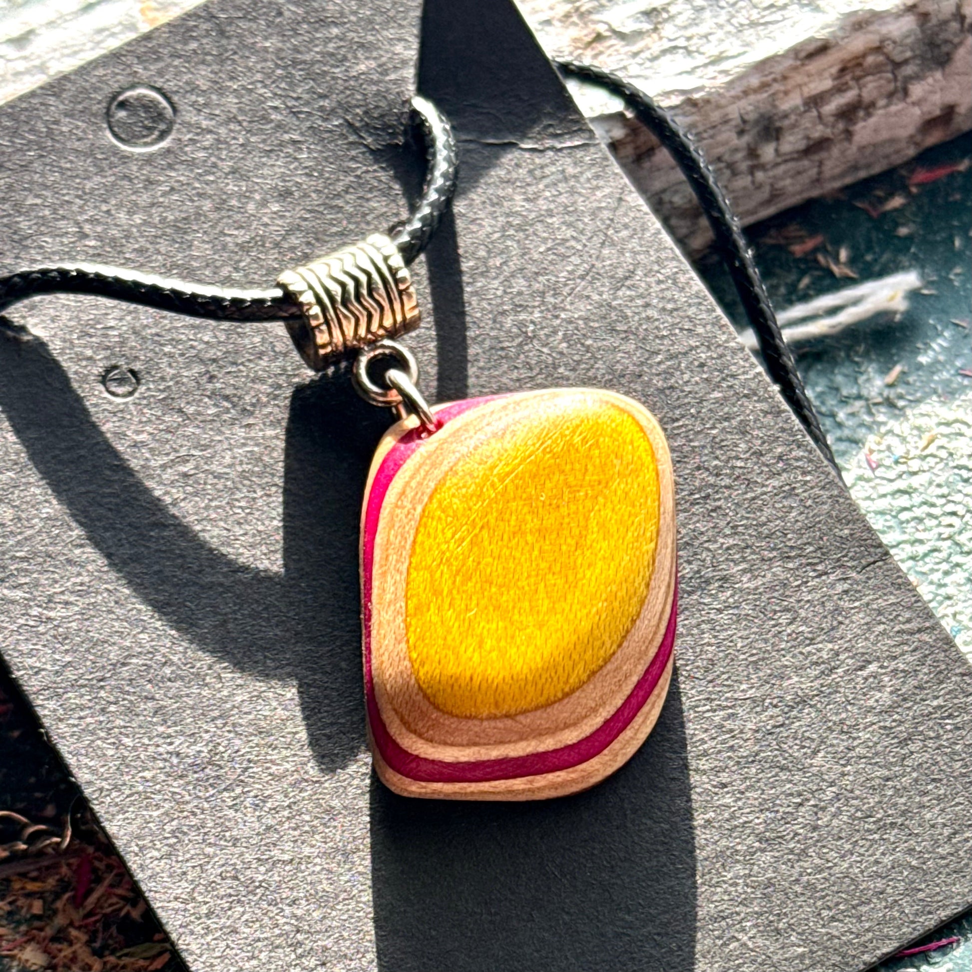 Yellow and pink layered wooden pendant carved from recycled skateboard maple on a black waxed cord.