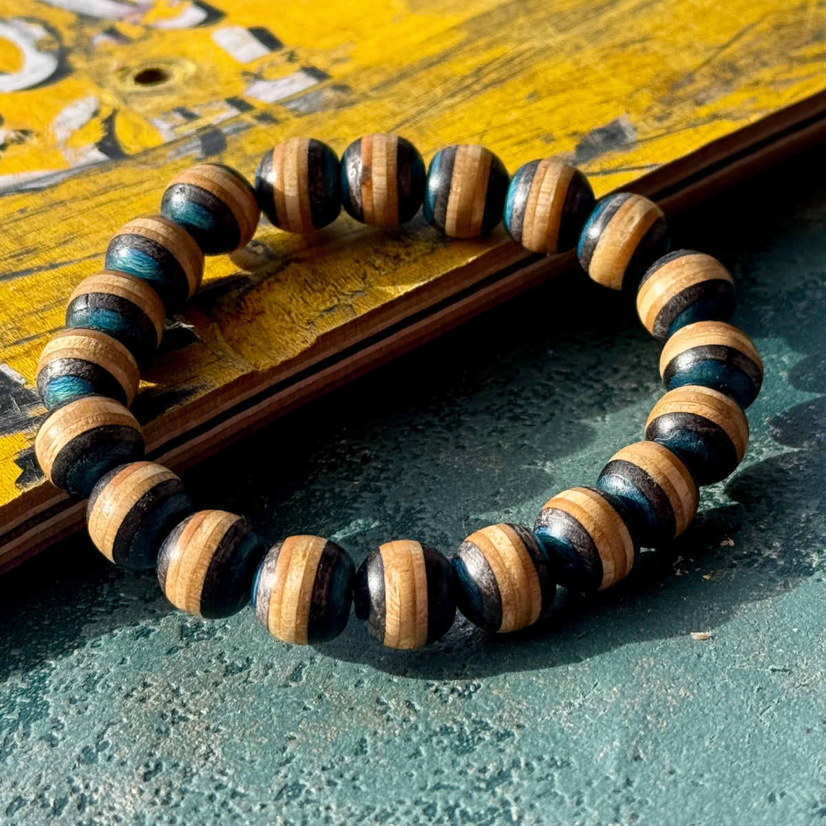 Blue and black recycled skateboard bracelet resting against a yellow deck with printed graphics, handmade by Baltic Dew.