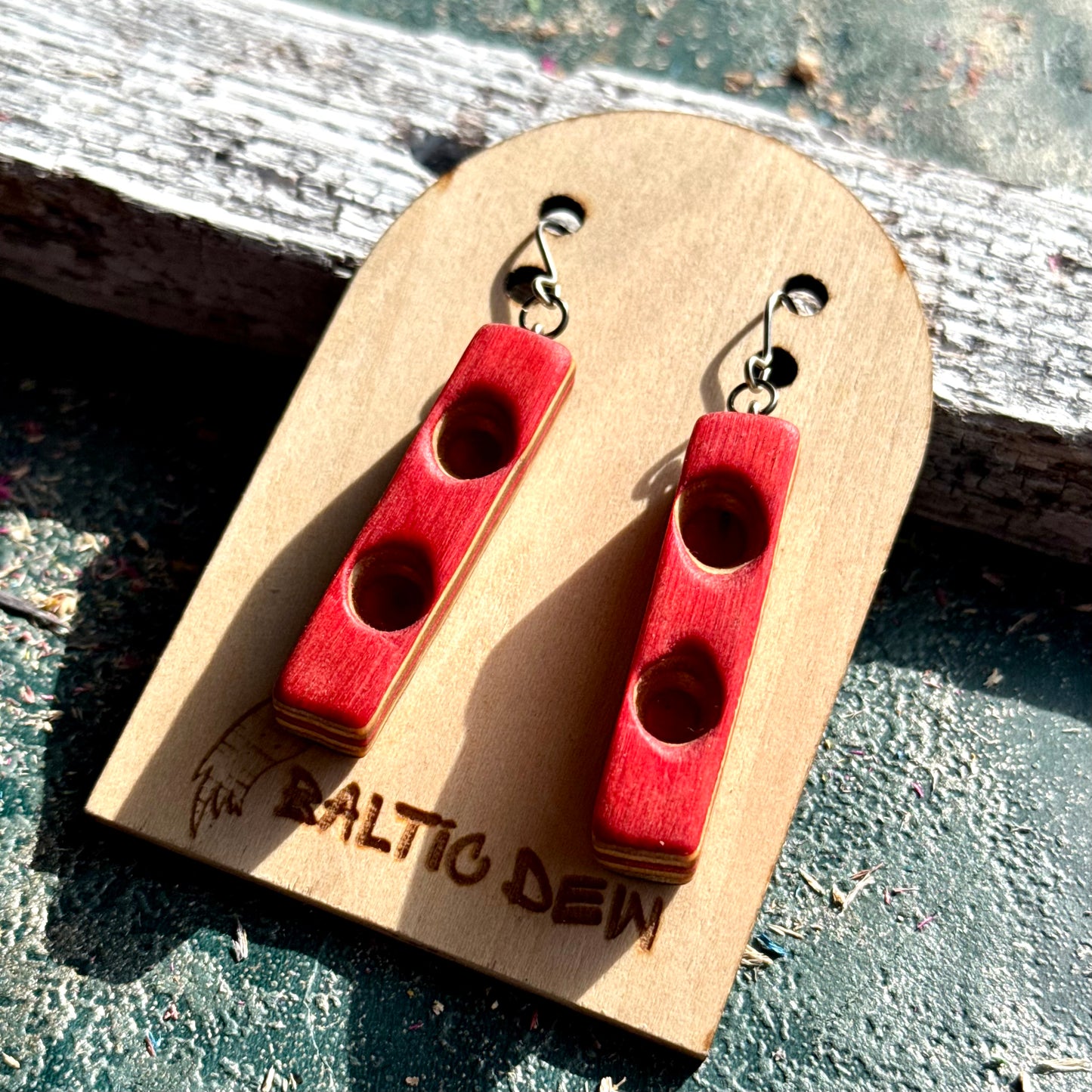 Red carved bar earrings made from recycled skateboard maple, shown on a Baltic Dew card outdoors