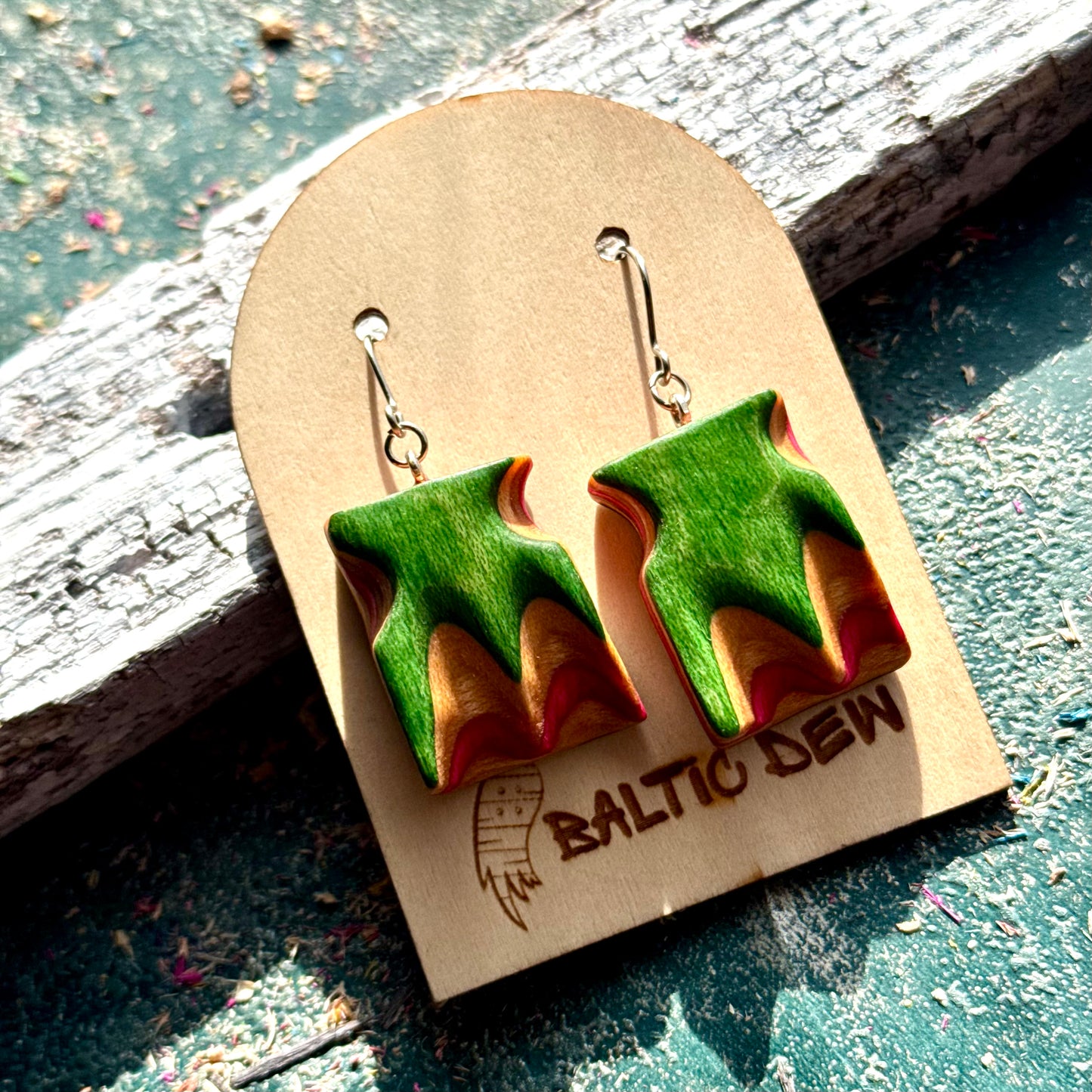Carved wave earrings made from recycled skateboard maple, shown on a Baltic Dew card outdoors
