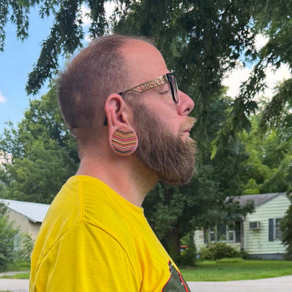 Bearded man wearing a striped wooden ear plug in a stretched lobe, photographed outdoors.