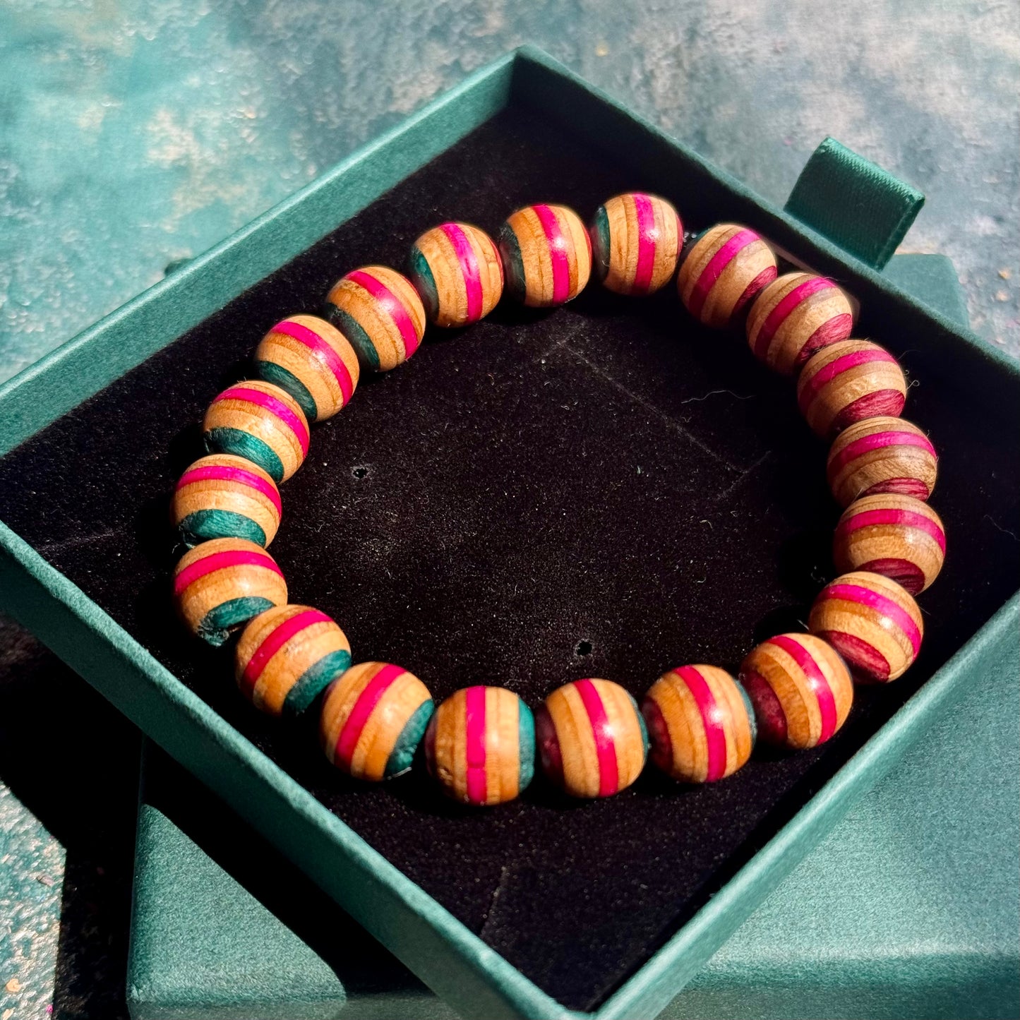 Pink and Teal Skateboard Bracelet made of round beads with alternating pink and teal stripes, displayed in a green jewelry box on a textured surface.