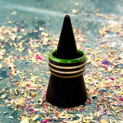 Green and black recycled skateboard wood ring with black stripe accents, displayed on a black cone stand over colourful wood shavings.