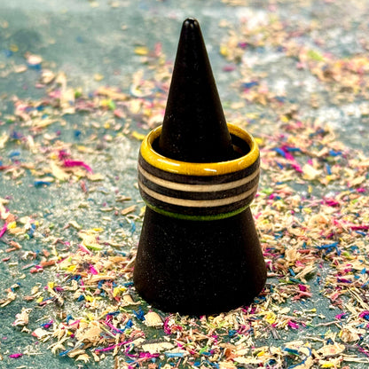 Yellow, green and black recycled skateboard wood ring with black and natural stripes, shown on a black cone stand surrounded by colourful wood shavings.