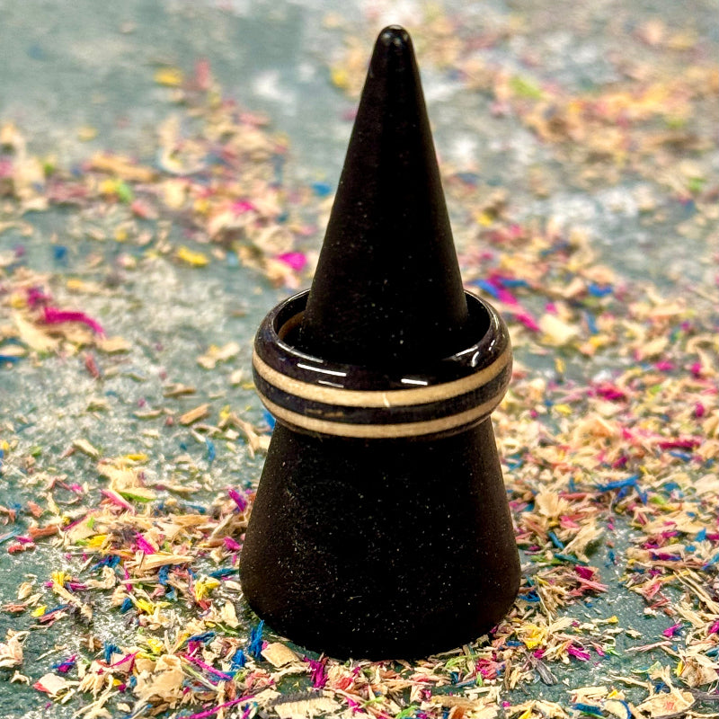 Single recycled skateboard wood ring in black and natural tones, displayed on a black cone stand with colourful wood shavings around it.