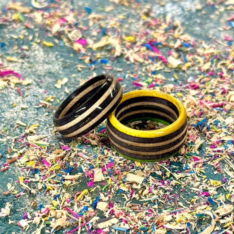 Two handmade rings from recycled skateboard wood in black, natural and yellow tones, displayed on a textured background.