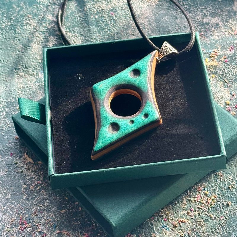 Teal curved skateboard pendant with geometric cutouts, displayed in a green jewellery box on a textured surface.