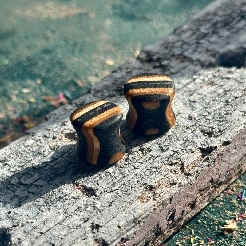 Black Double Flare Wooden Ear Plugs 10mm with striped natural wood pattern resting on weathered wood surface outdoors.