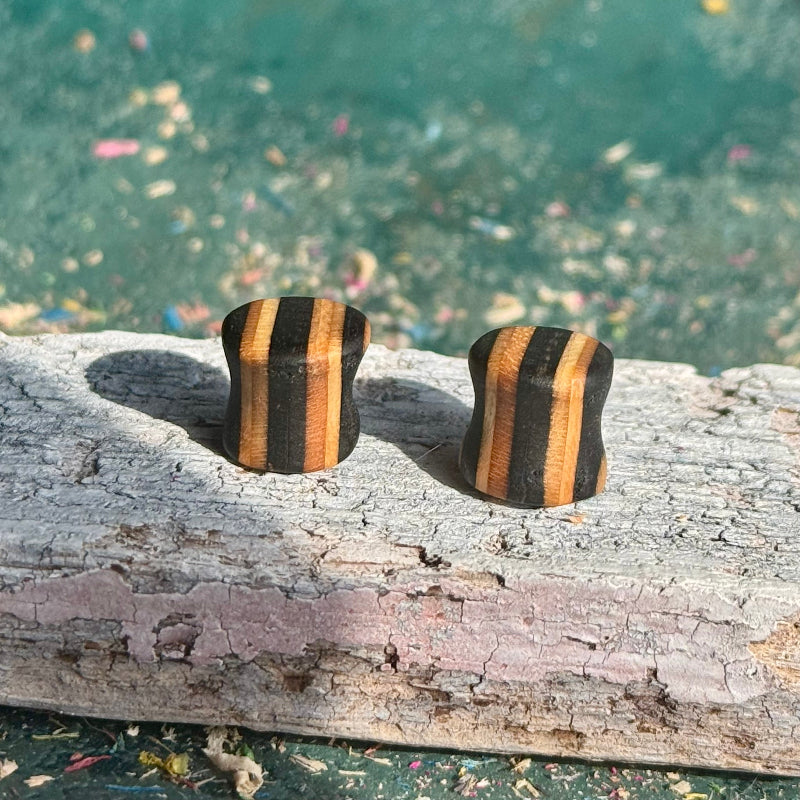 Black Double Flare Wooden Ear Plugs 10mm with striped natural wood design placed on weathered wood surface outdoors