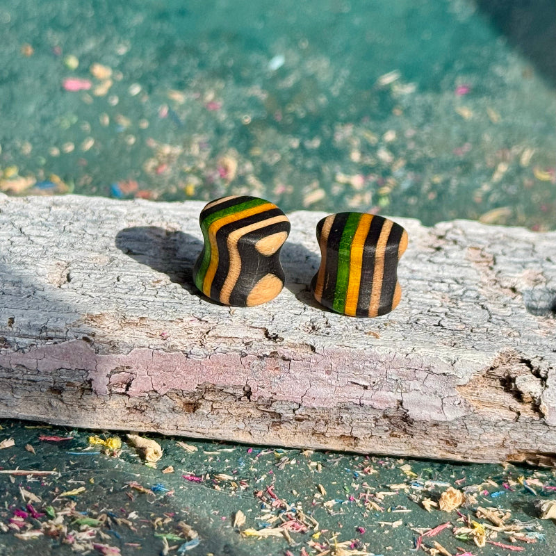 Green, yellow and black 10mm ear plugs in recycled skateboard wood, resting on a weathered plank.
