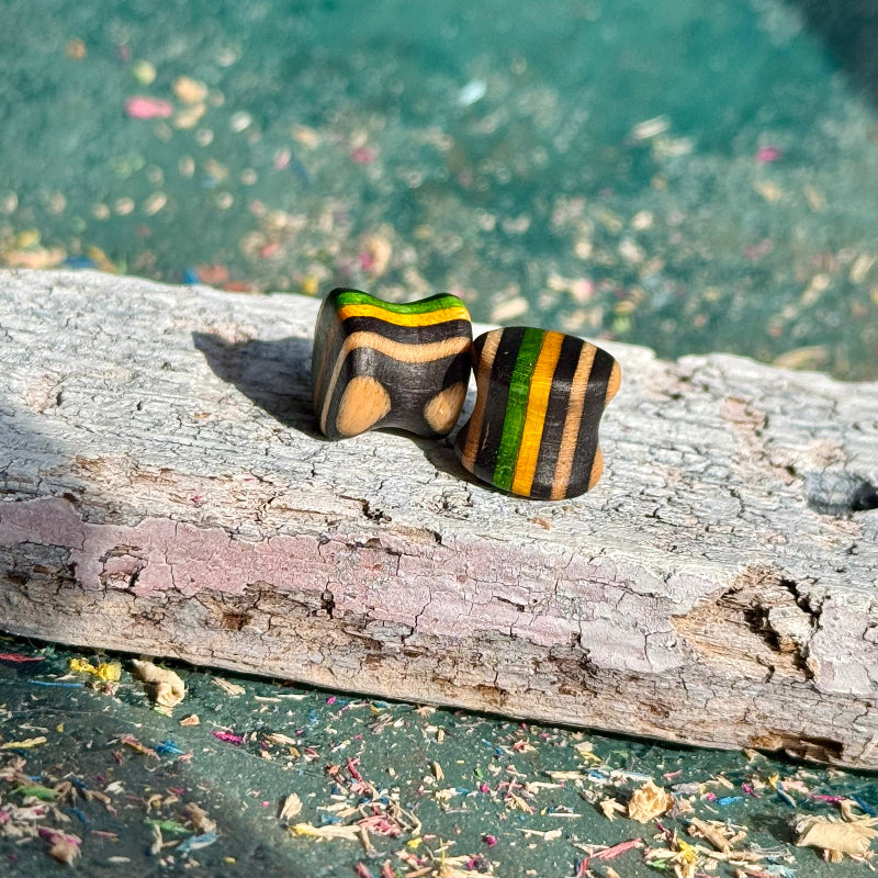 Green, Yellow and Black Double Flare Wooden Ear Plugs 10mm resting on weathered wood with blurred colorful background.