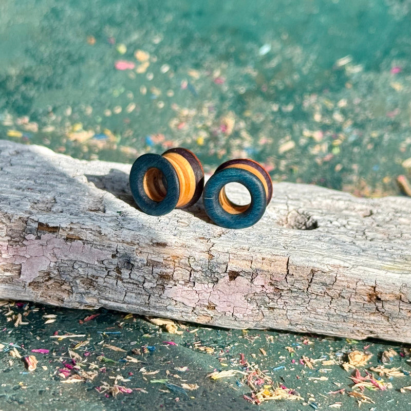 Orange, Purple and Blue Double Flare Wooden Ear Tunnels 10mm resting on weathered driftwood with scattered flower petals.