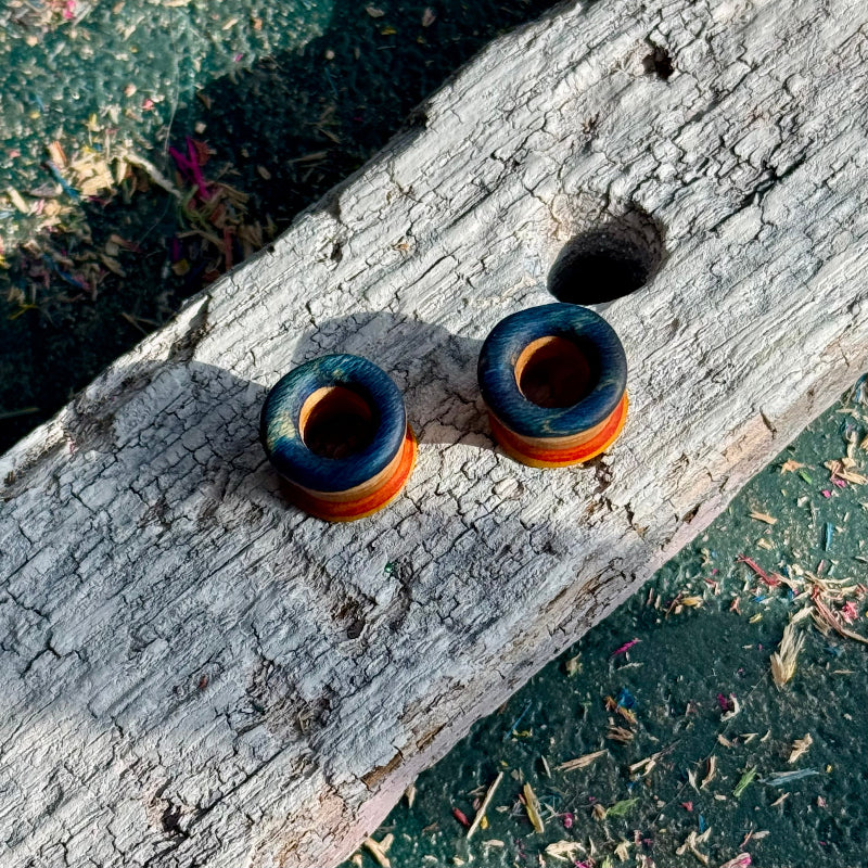 Yellow, Orange and Blue Double Flare Wooden Ear Tunnels 10mm placed on textured weathered wood outdoors.