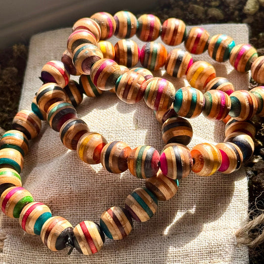 A stack of colorful beaded bracelets made from recycled skateboard wood, each featuring unique striped patterns in vibrant hues of pink, green, blue, and natural wood tones, displayed on a textured beige cloth in natural sunlight.