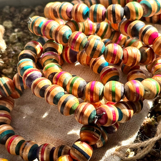 A close-up of multiple colorful beaded bracelets made from recycled skateboard wood, featuring vibrant striped patterns in pink, green, blue, orange, and natural wood tones, coiled together on a beige fabric surface in warm sunlight.