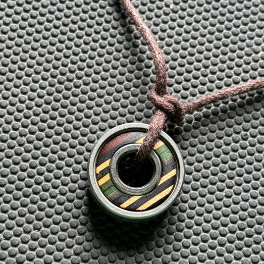 Close-up of a handmade necklace featuring a recycled skateboard wooden pendant encased in a metal ball bearing, strung on a brown cord against a textured black background.