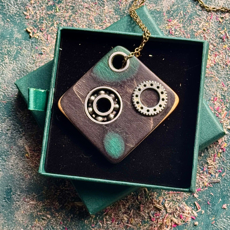 Square skateboard maple pendant with a steel bearing and toothed gear, on a brass chain in a green box.